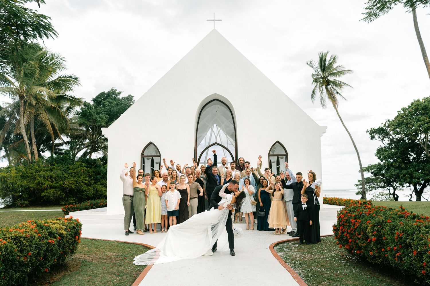 Bride and groom kiss outside a white tropical chapel surrounded by cheering wedding party and palm trees.