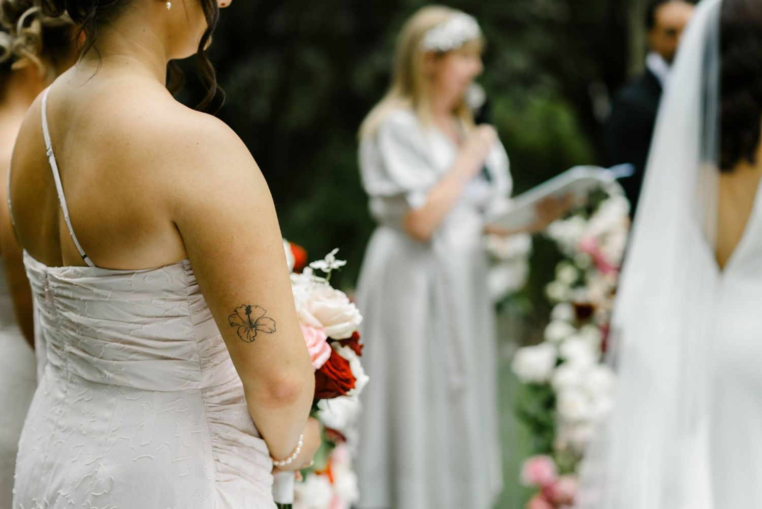 Bridesmaids in elegant silver satin dresses hold bouquets during an outdoor wedding ceremony.