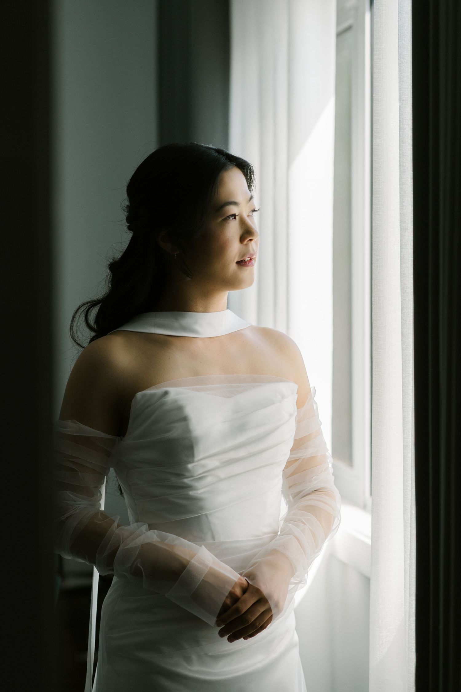 A bride in a white off-shoulder tulle gown gazes out a window in soft natural light.