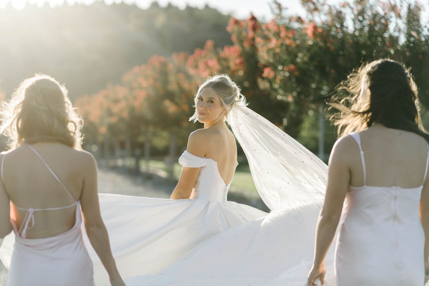 Bride in off-shoulder white gown and veil walks with bridesmaids in white dresses outdoors in golden sunlight.