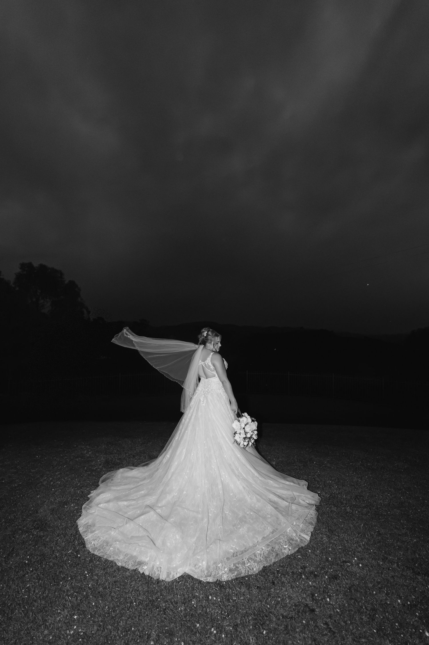 Bride in flowing white gown and veil blowing in wind, holding bouquet, standing outdoors at night in black and white photo.