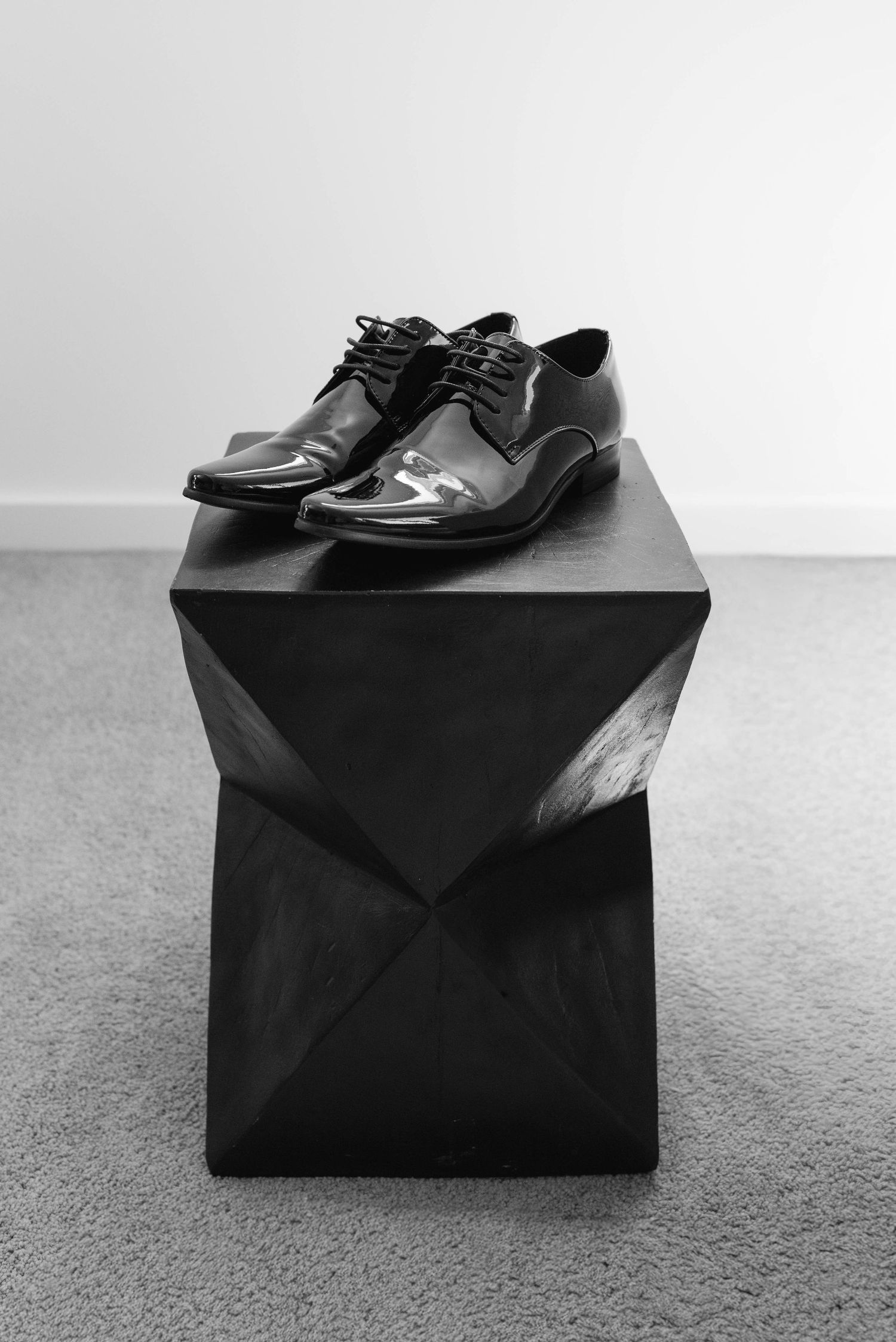 Shiny black patent leather oxford dress shoes displayed on a geometric dark wooden pedestal in black and white.