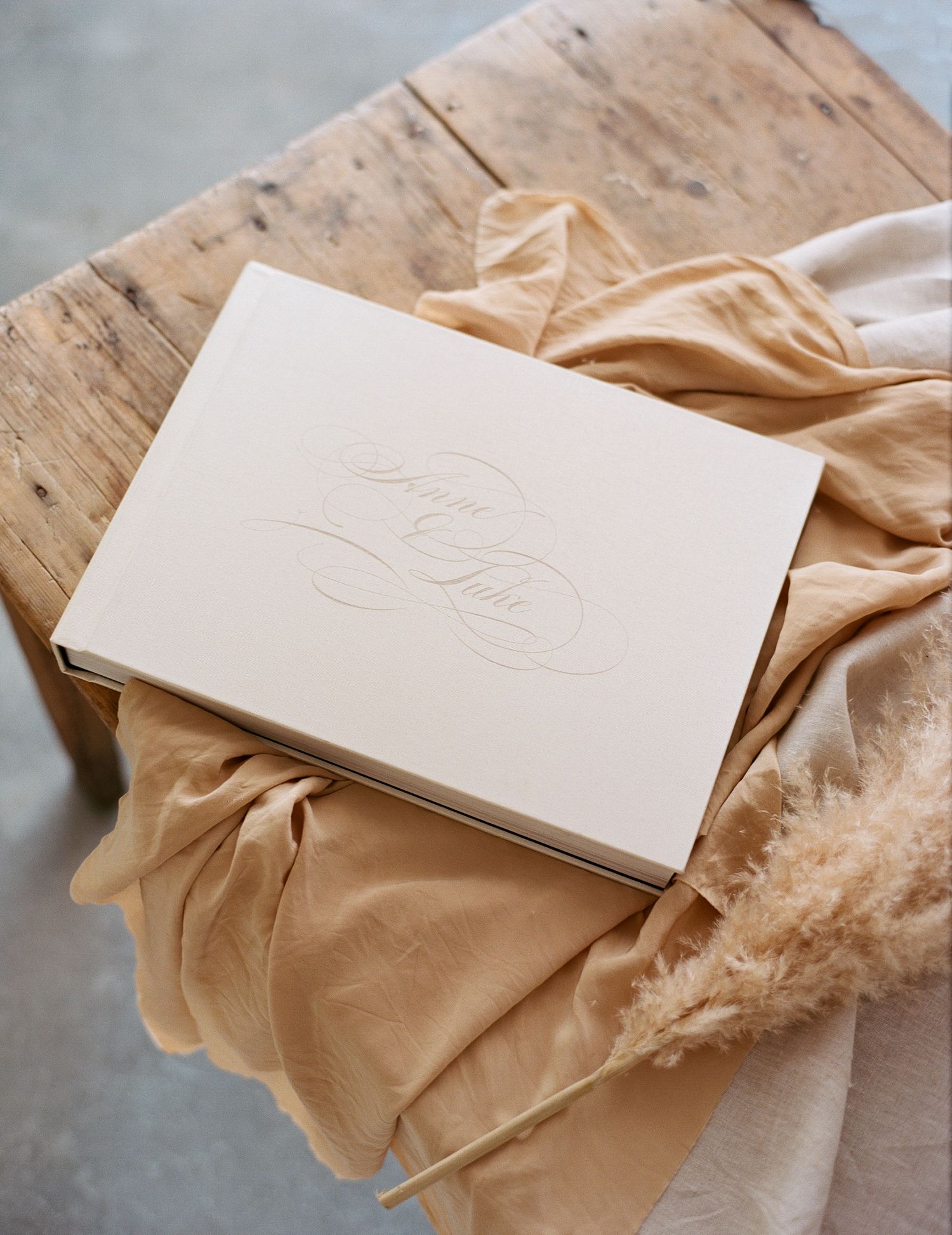 Elegant cream embossed wedding guest book resting on rustic wood table with golden silk fabric and pampas grass.