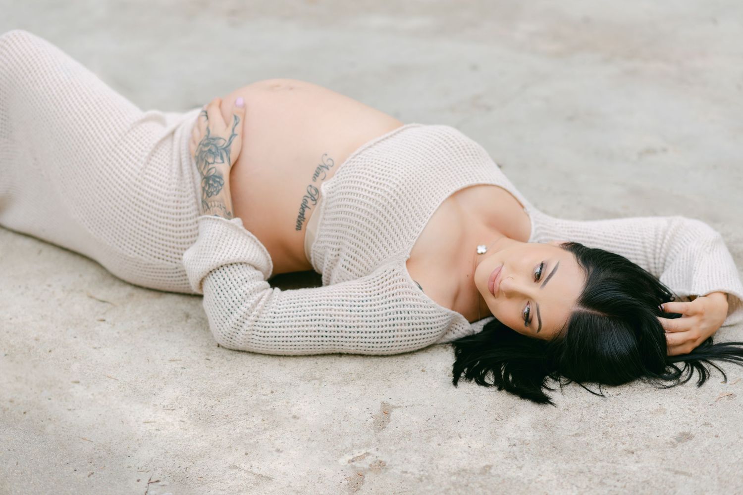 Pregnant woman with tattoos lying on ground wearing cream knit crop top, showcasing baby bump in outdoor maternity photo.