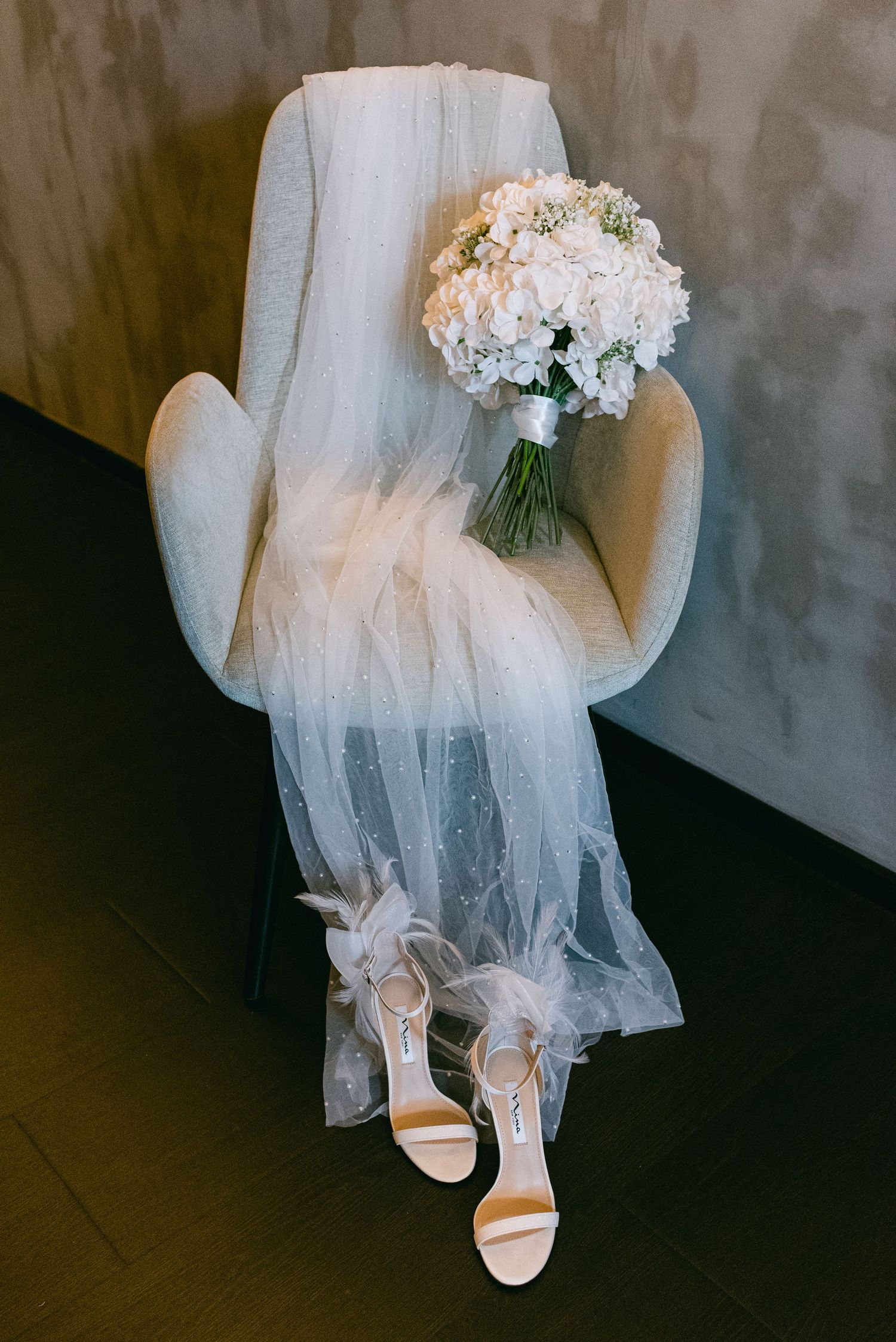 A wedding veil and bouquet of white hydrangeas rest on a cream armchair with white heels placed beneath.