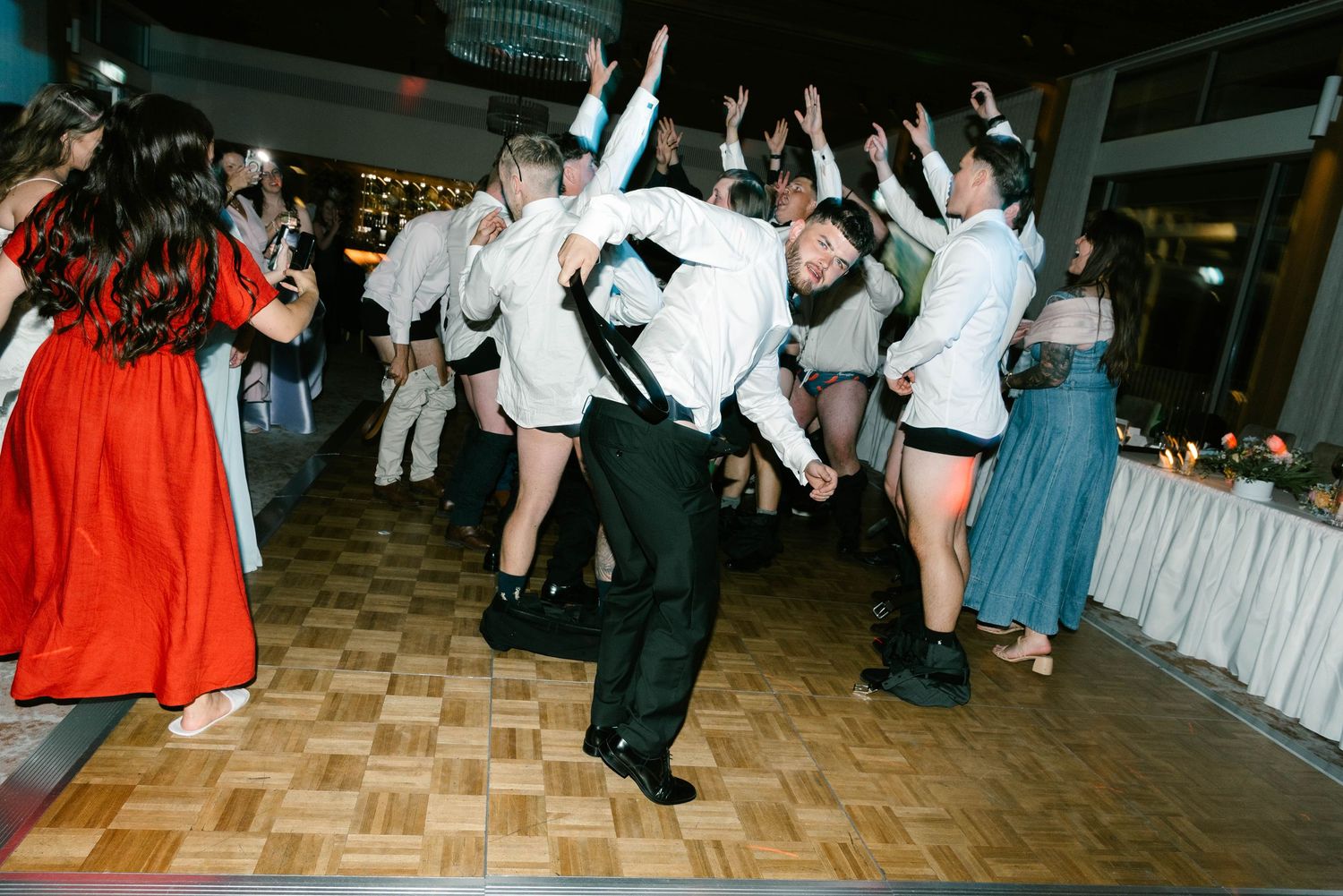 Guests in formal attire dancing and jumping enthusiastically on a parquet dance floor at a wedding celebration.