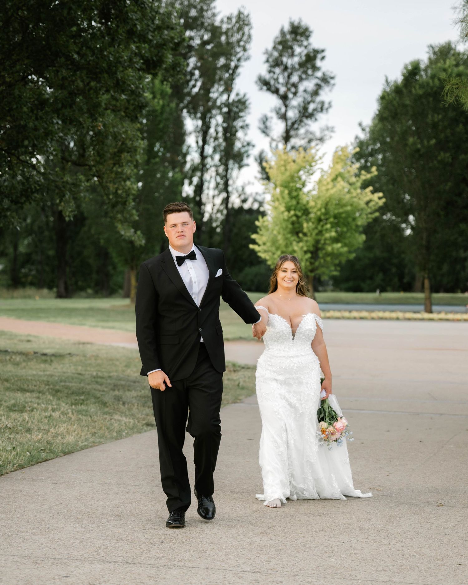A couple in formal wedding attire walks along a tree-lined path during their outdoor wedding photoshoot.