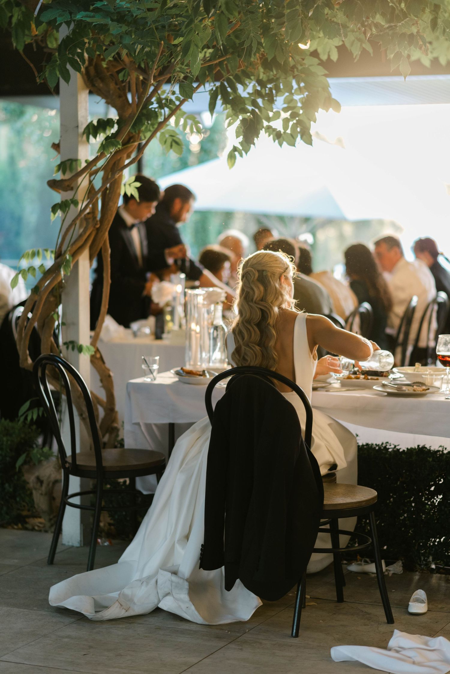 Bride in white gown seated at elegantly decorated outdoor wedding reception table beneath lush green tree.
