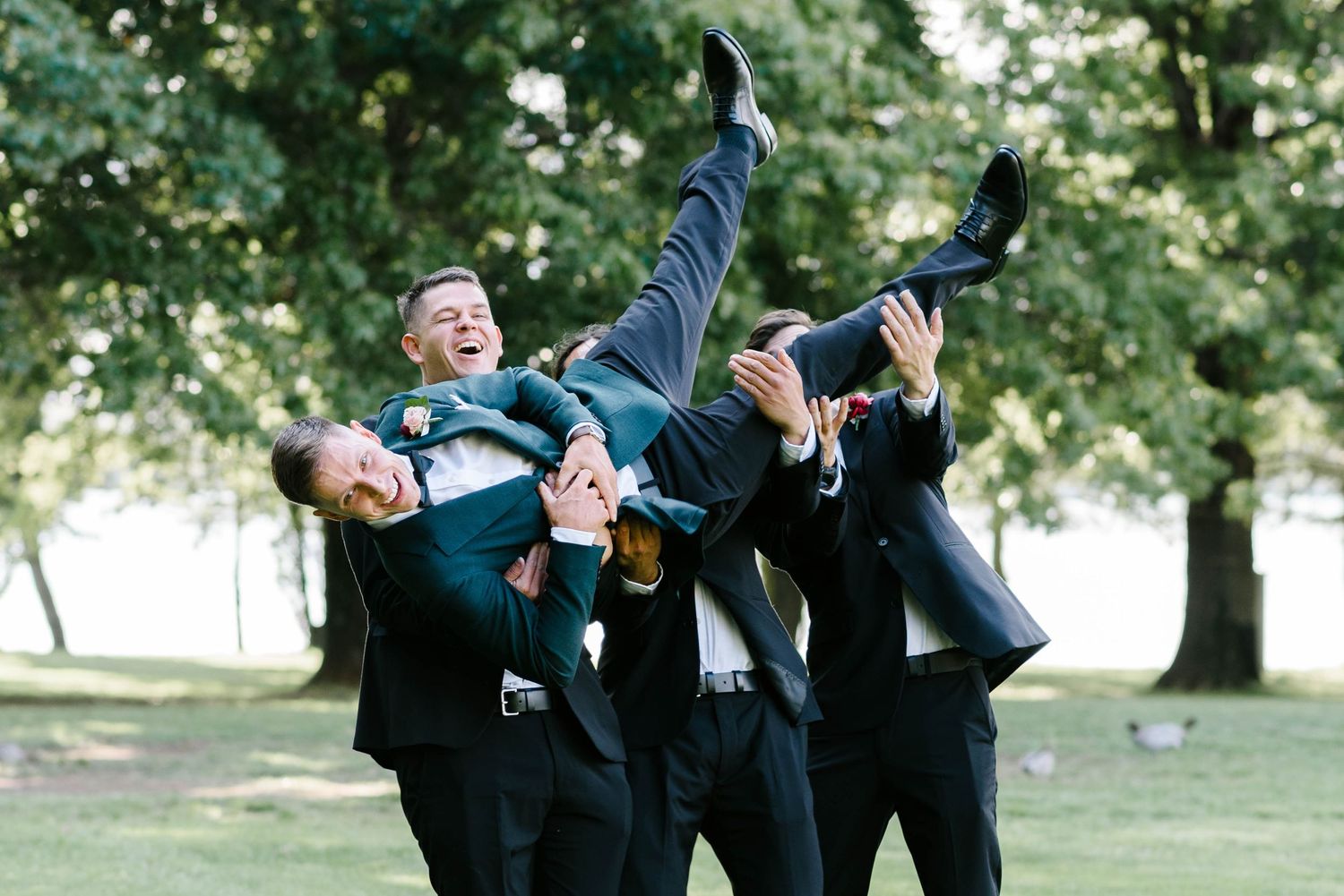 Groomsmen in dark suits strike playful poses while celebrating outdoors in a sunny park setting.