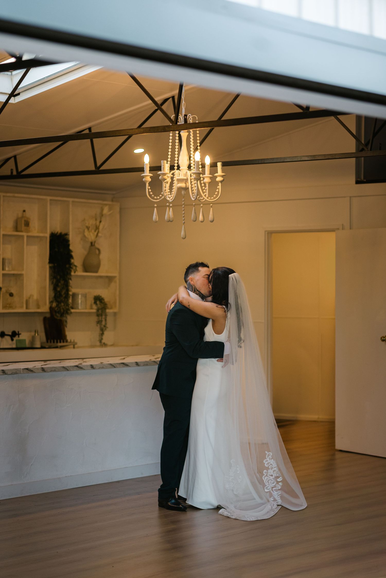 Bride and groom share first kiss under chandelier in elegant indoor wedding venue.
