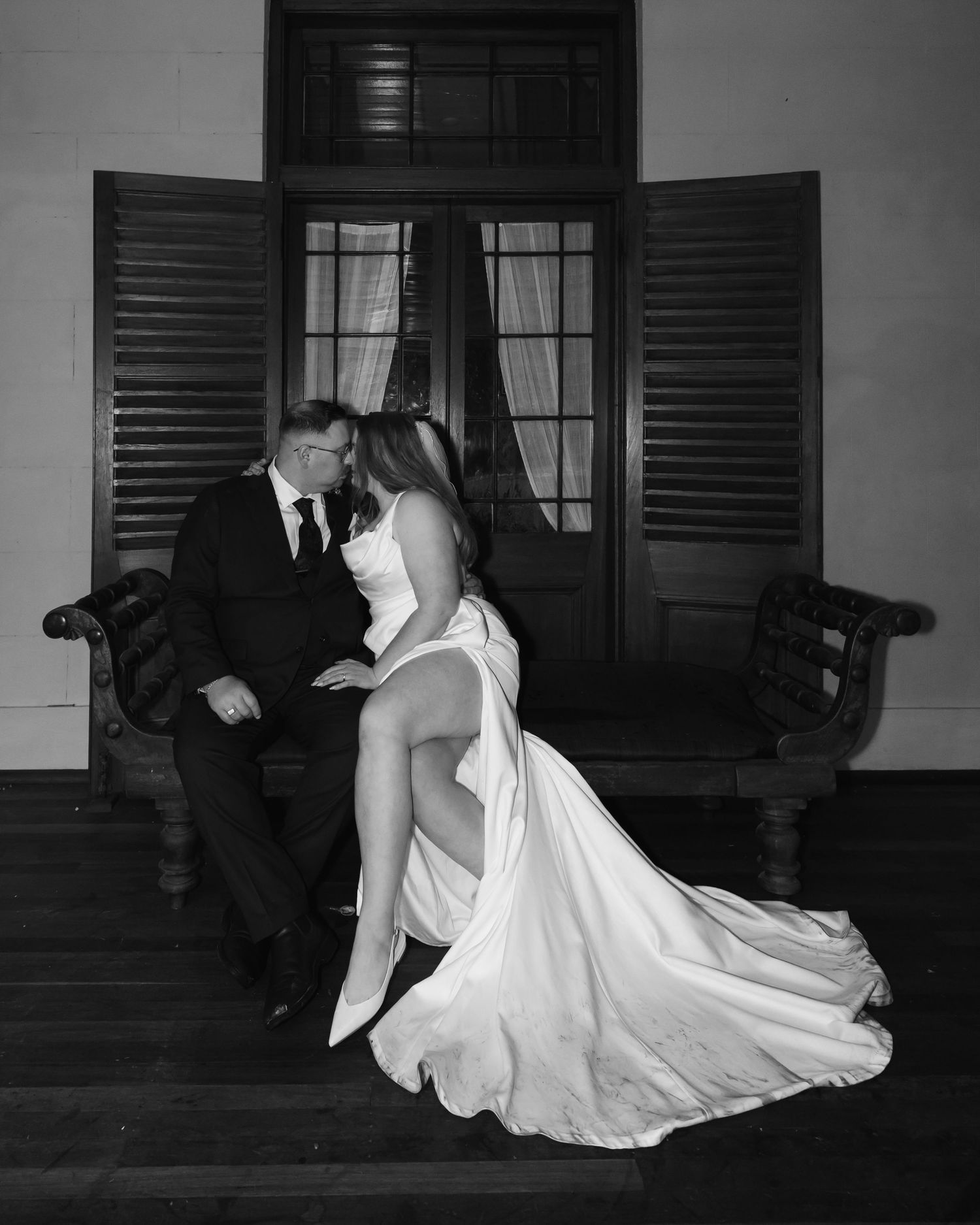 Black and white photo of a bride and groom sharing an intimate moment on a bench beside shuttered windows.