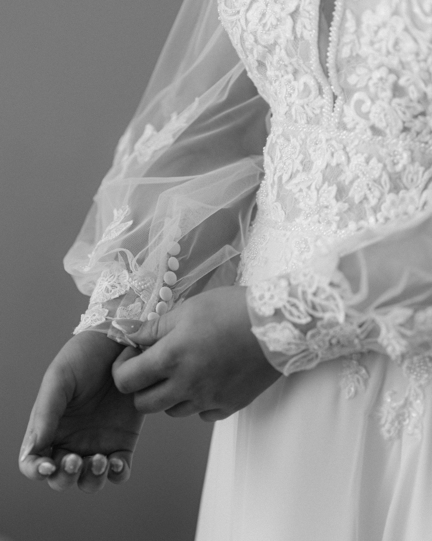 Close-up black and white photo of a bride in lace wedding gown with sheer sleeves, hands clasped together.