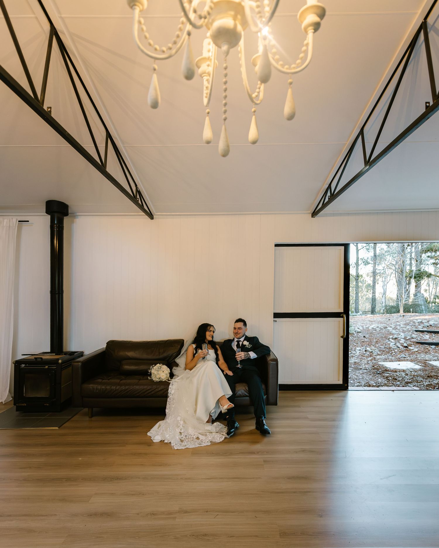 Bride and groom sit together on a leather sofa beneath a chandelier in a modern white venue with open glass doors.
