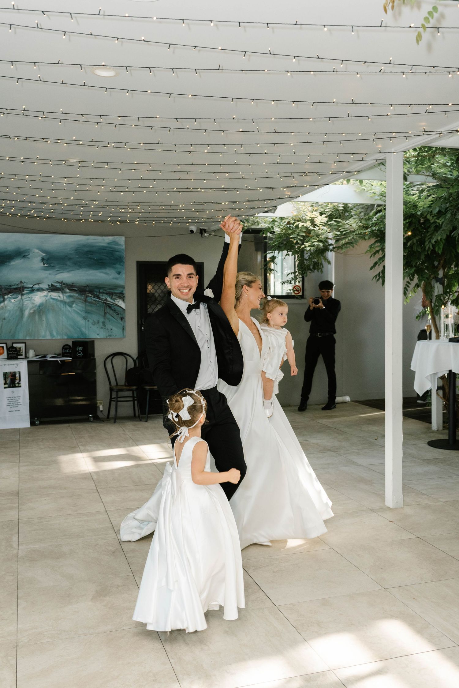 Bride and groom dancing with flower girls under fairy lights at elegant wedding reception venue.