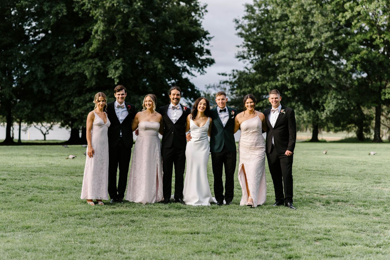 Wedding party in formal attire poses together on a green lawn with trees in the background.