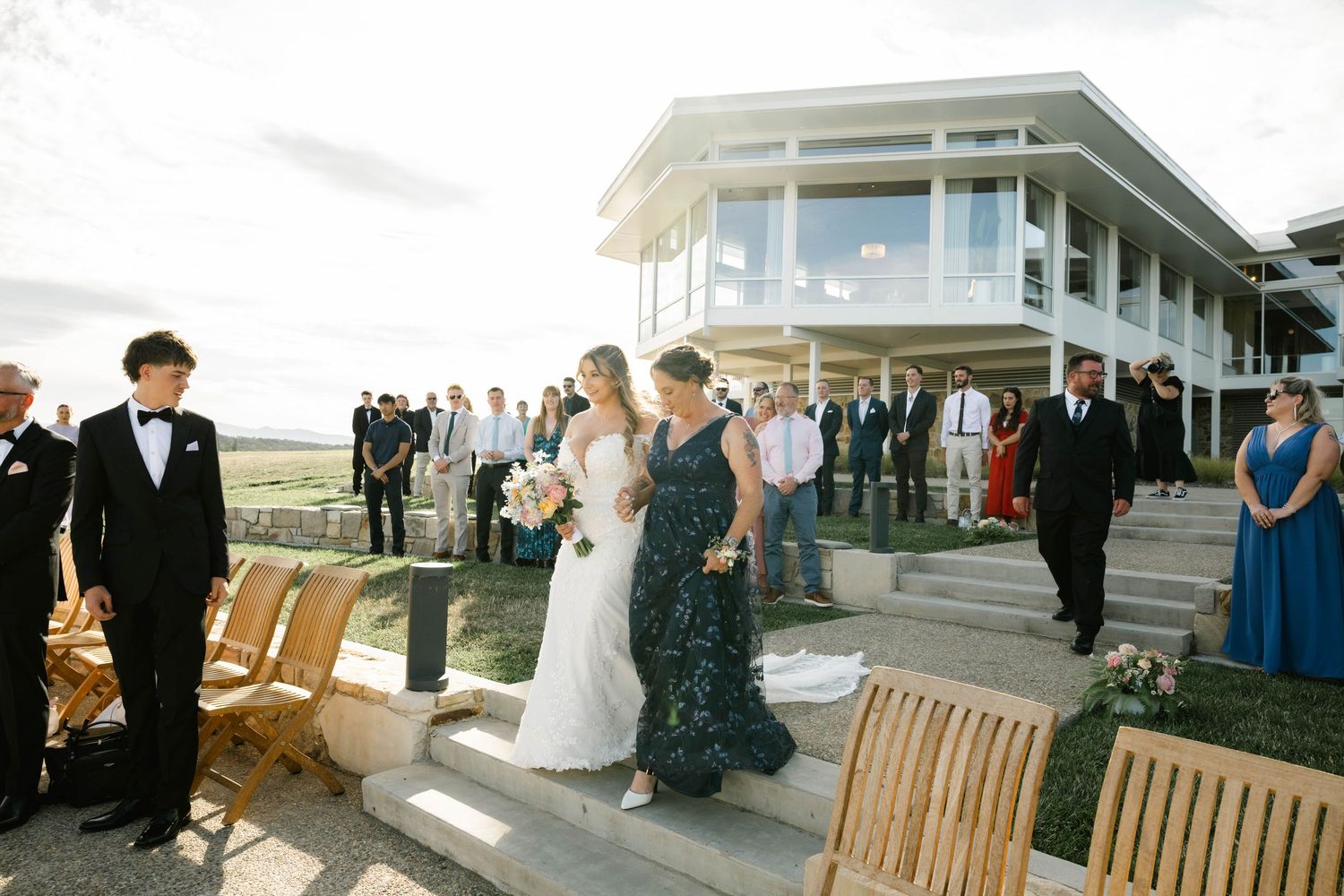 Wedding ceremony guests seated in wooden chairs on a waterfront deck overlooking a scenic coastal view.