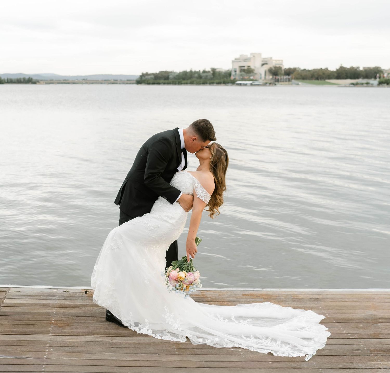 A romantic wedding photo on a dock with a lake and building in the background.