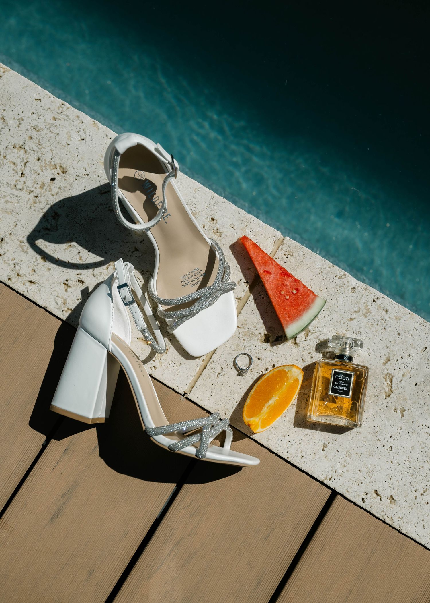 White sandals and perfume bottles arranged artistically by a turquoise pool edge.