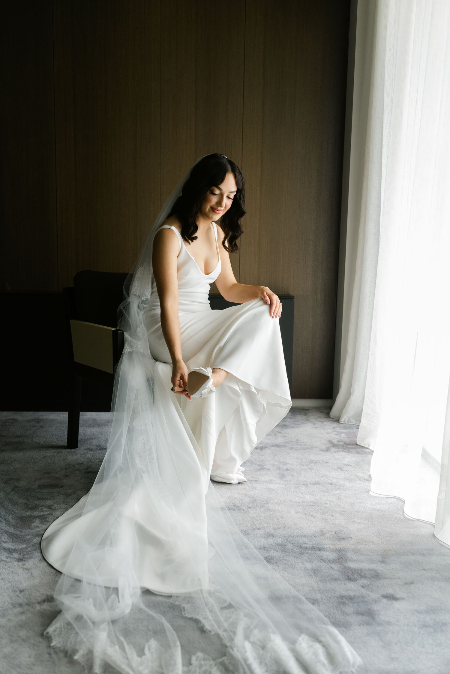 A bride in a white gown and veil sits by a window, smiling as she adjusts her shoe before her wedding.