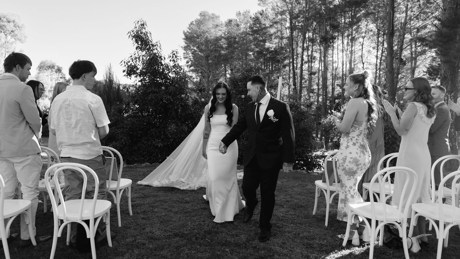 Black and white photo of a bride and groom walking down the aisle at an outdoor wedding ceremony.