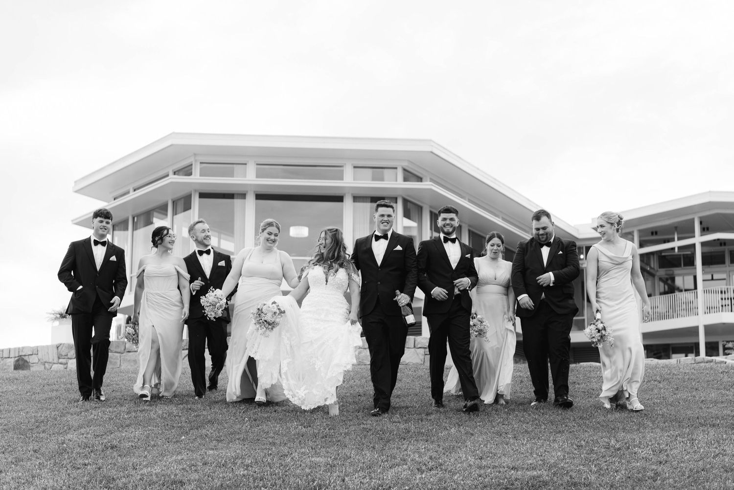 Wedding party in formal attire walks and poses together on a grassy lawn in front of a modern building.