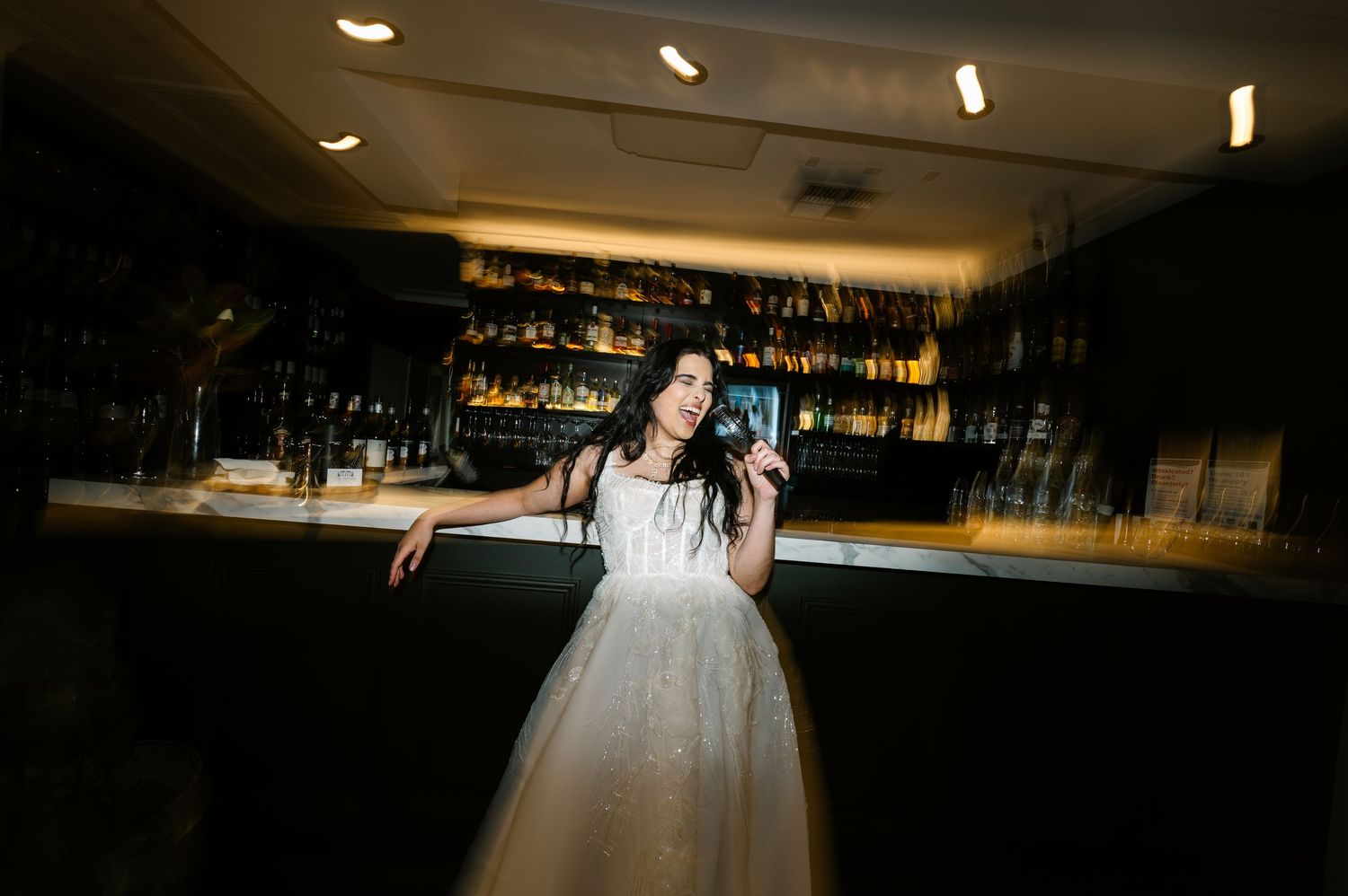 Woman in white tulle gown holding a drink at a dimly lit bar with bottle-lined shelves.
