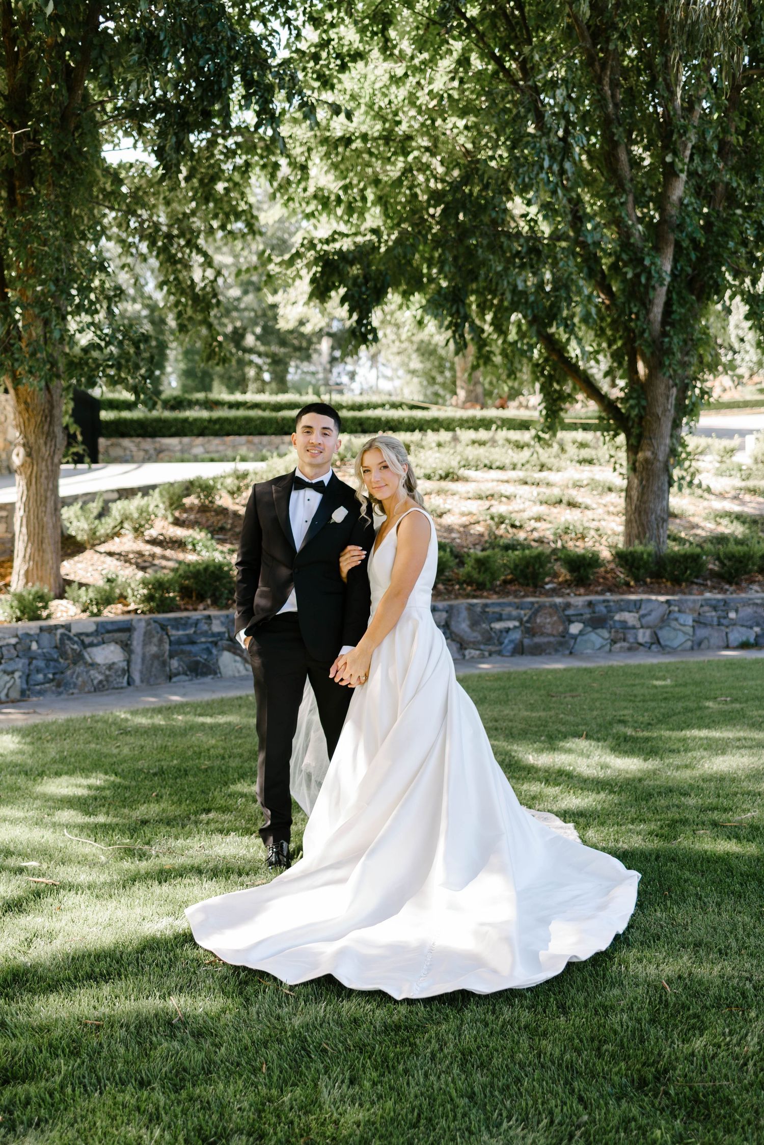 Bride and groom posing on green lawn with stone wall and trees at outdoor wedding venue.