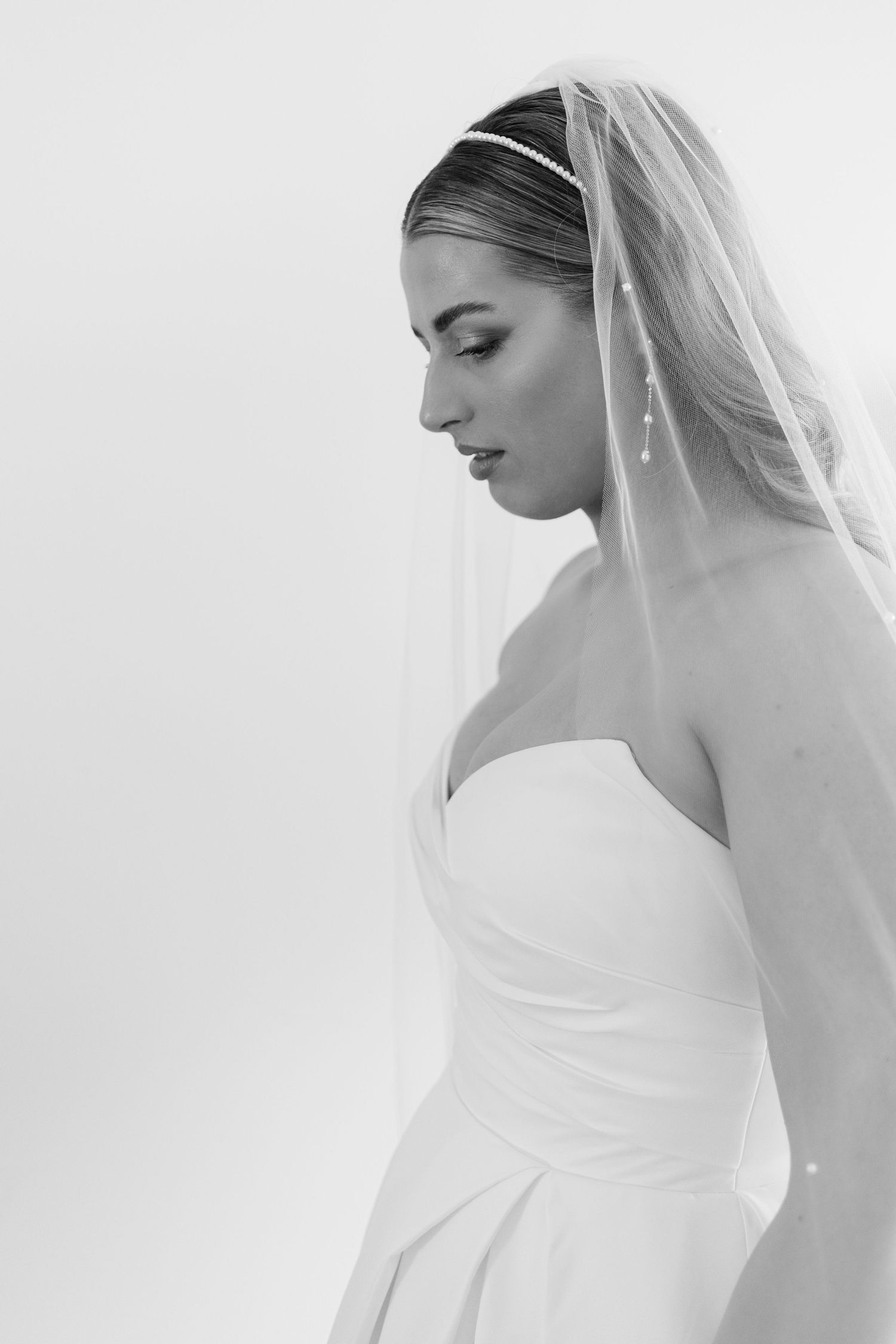 Black and white profile portrait of a bride in strapless gown with veil and headband, looking down elegantly.