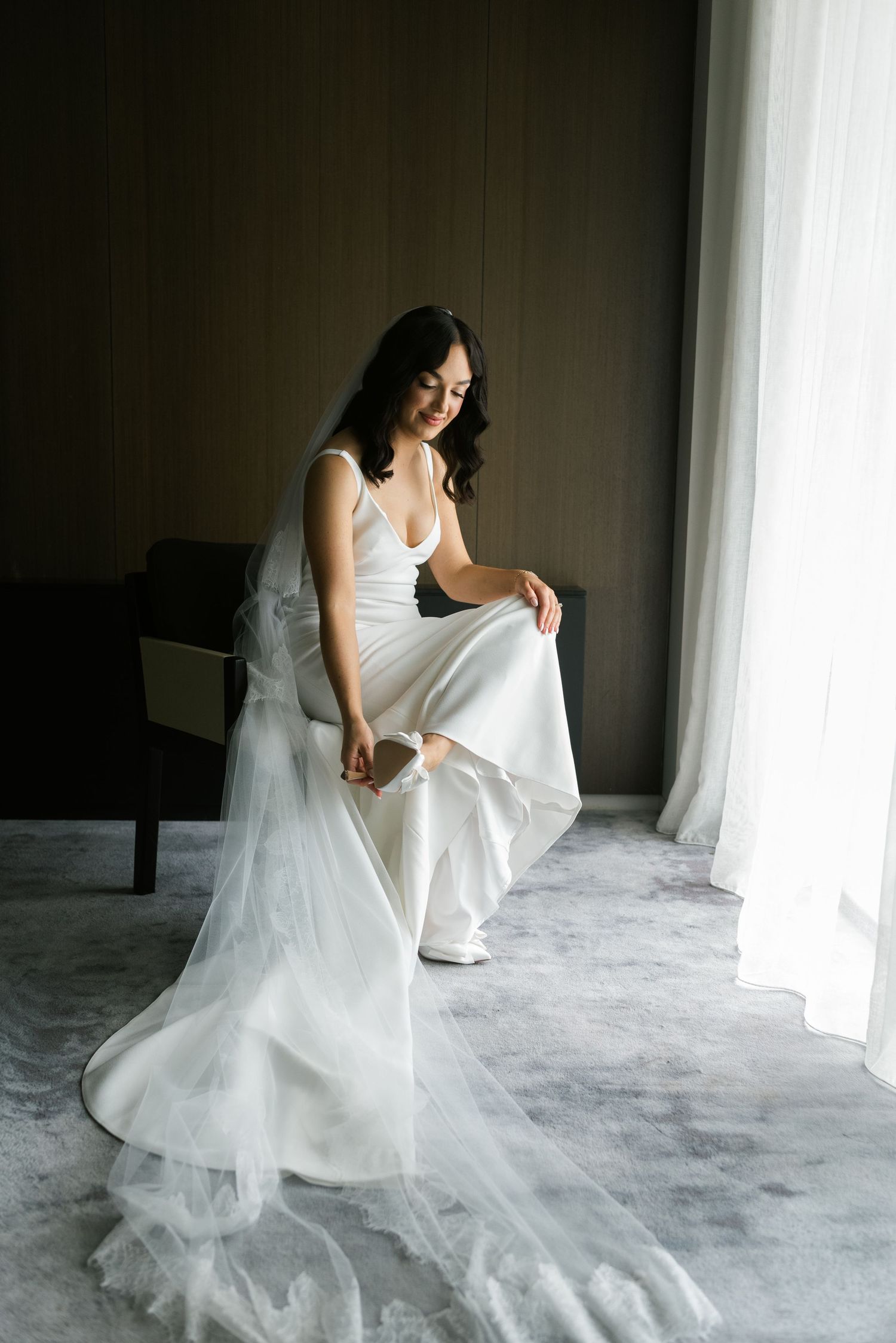 Bride in white wedding gown and veil sits by window, adjusting her shoe in an elegant hotel room.