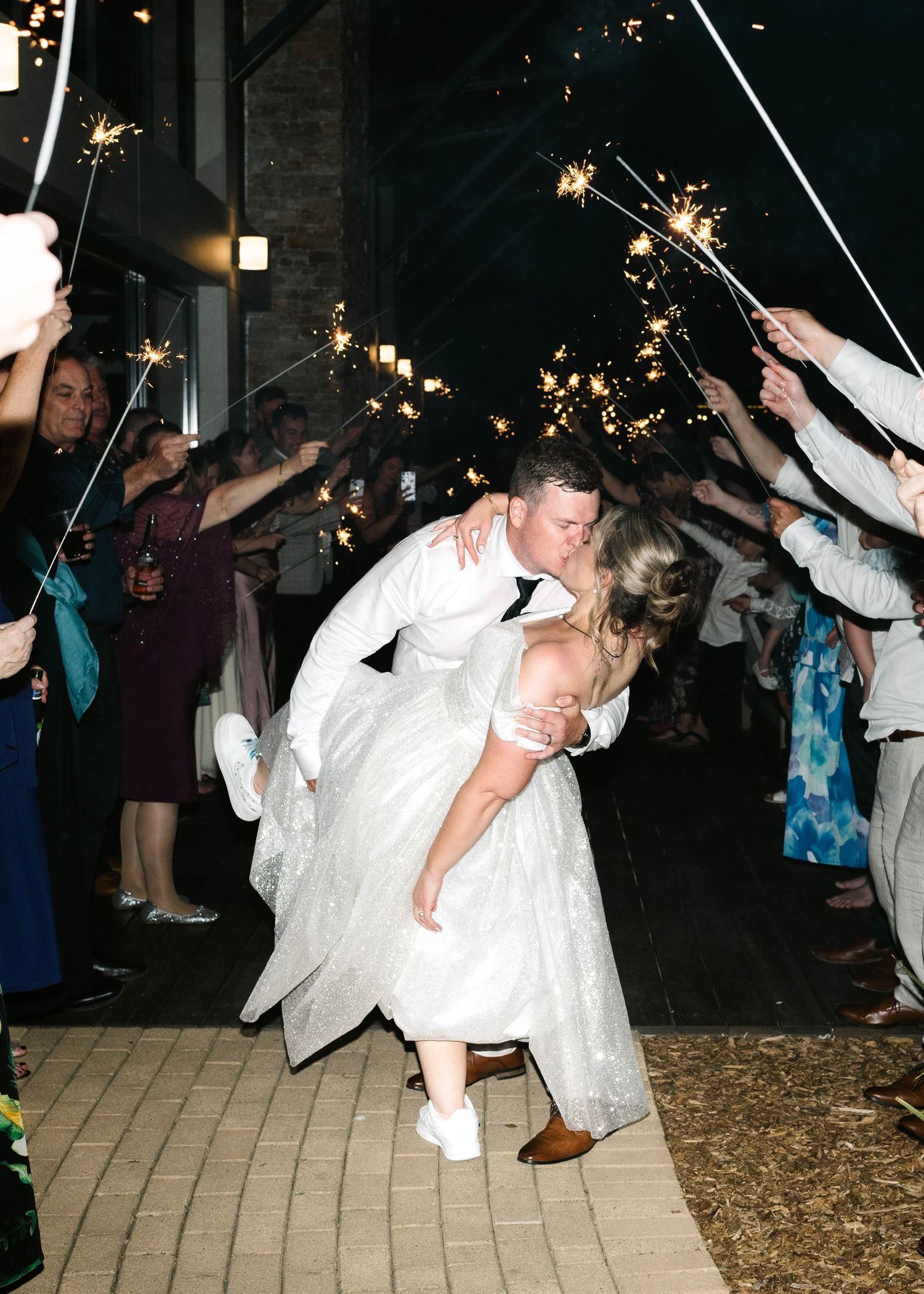 Newlywed couple shares romantic dip kiss under sparklers during wedding reception exit on brick pathway.
