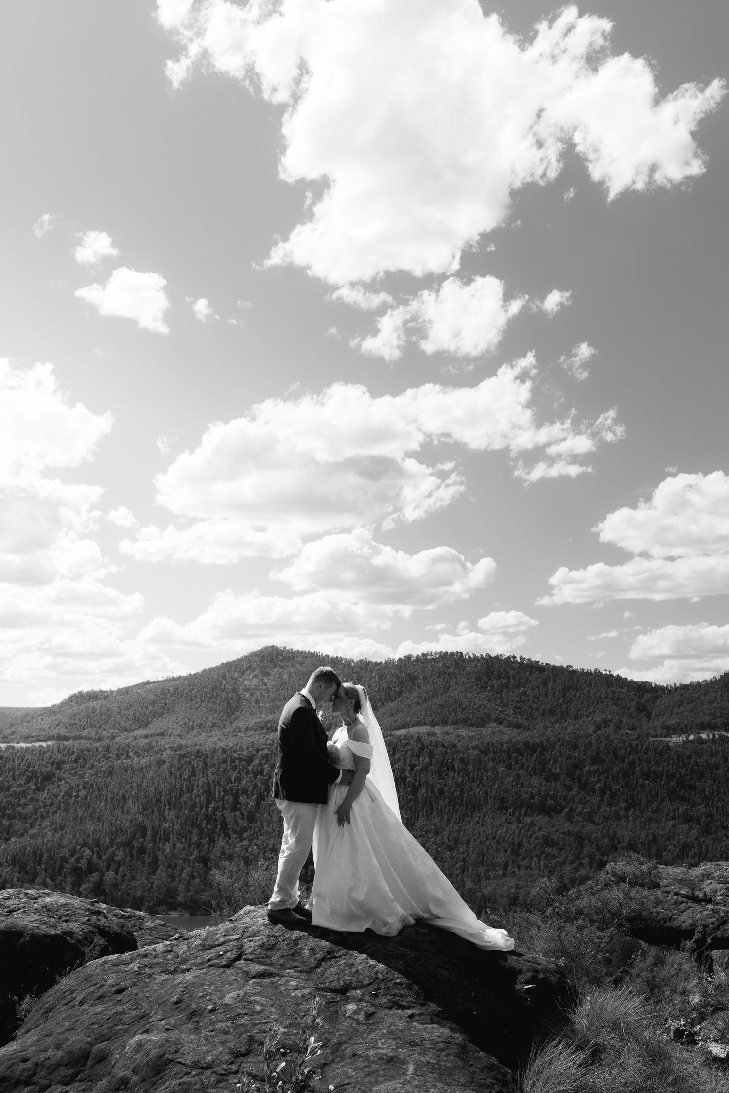 Black and white photo of bride and groom embracing on rocky summit with forested mountains and cloudy sky backdrop.