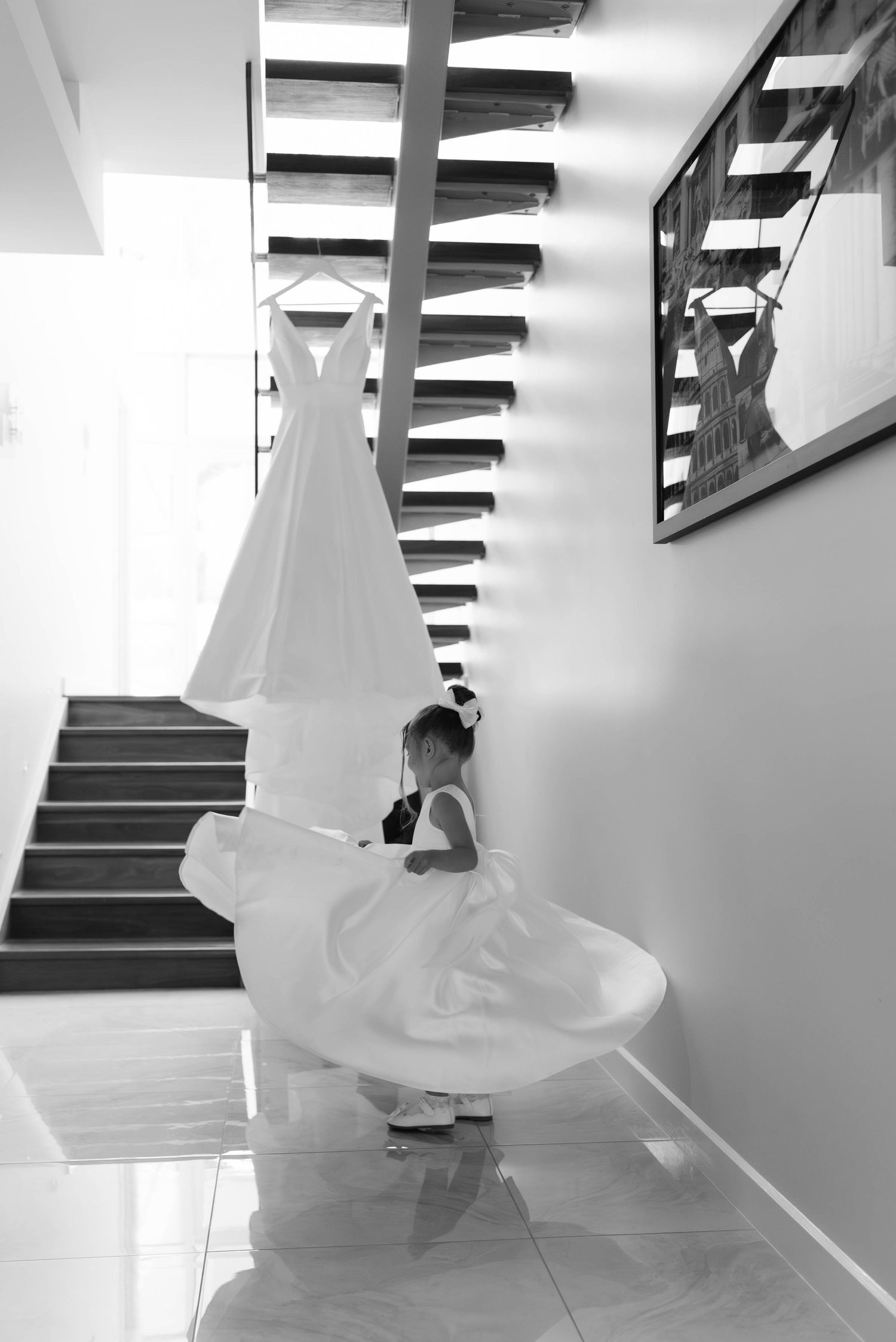 Young flower girl in white dress twirling near a bridal gown hanging on a staircase in black and white.