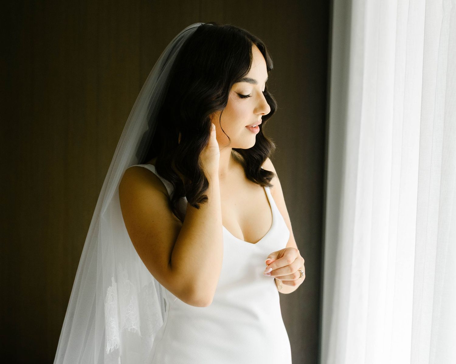 A bride in a white wedding dress and veil stands in soft window light with a serene expression.
