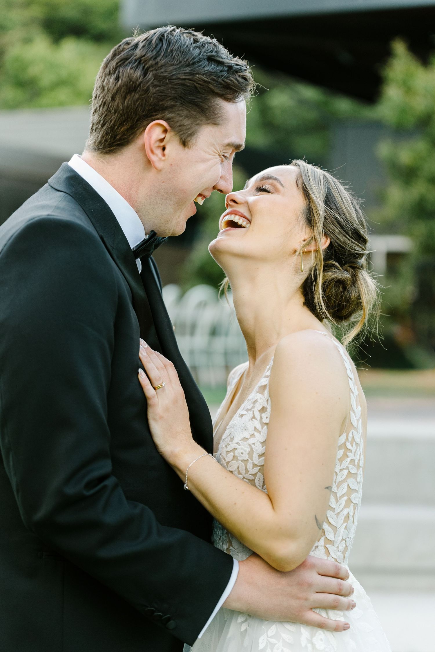A joyful bride in a floral gown laughs with her groom in a black tuxedo during their outdoor wedding celebration.