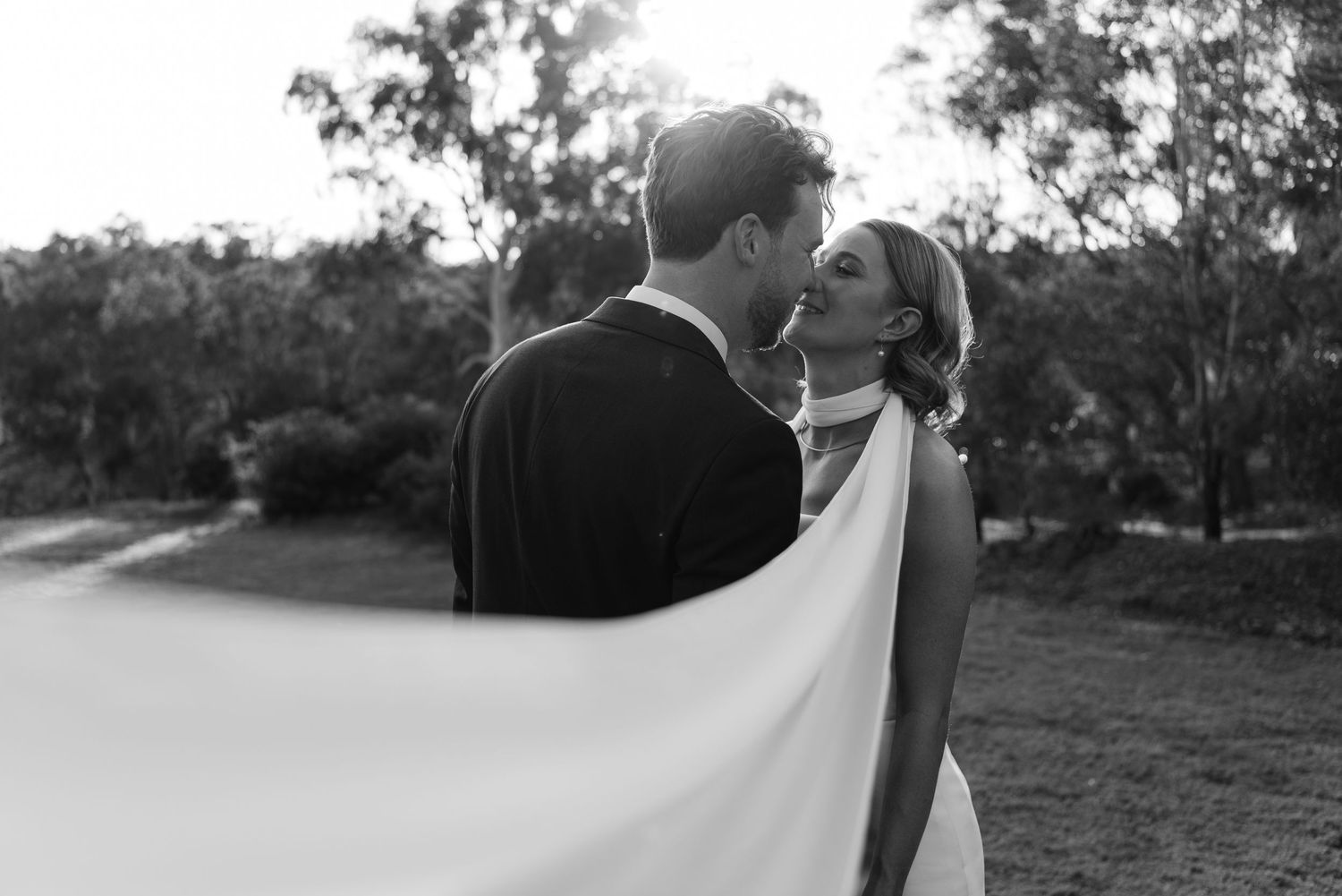 Black and white photo of a bride and groom sharing a romantic moment outdoors, with her veil flowing in the breeze.