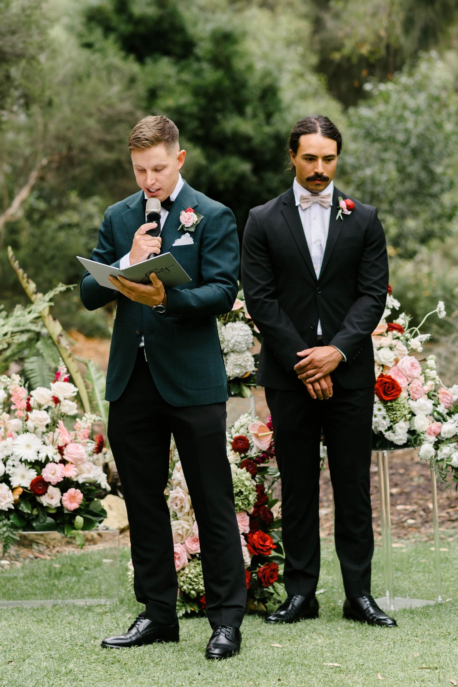 Young boys in formal black suits and white shirts reading from books during an outdoor wedding ceremony.