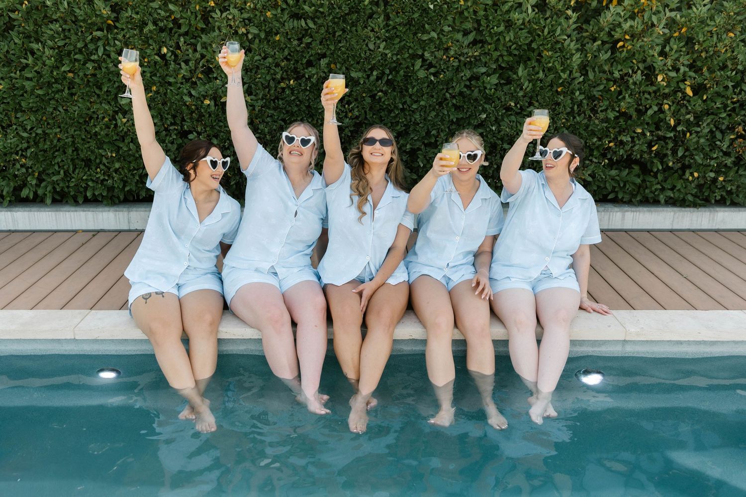 Five women in matching white outfits sit poolside raising champagne glasses, wearing fun sunglasses at a bachelorette party.