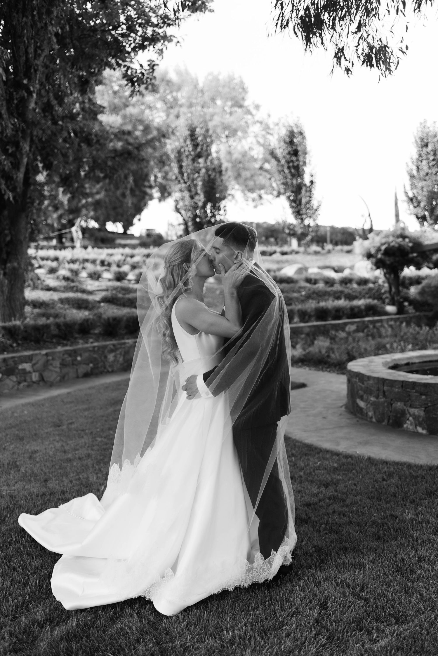 Black and white photo of a bride and groom kissing under a veil in a lush garden setting.