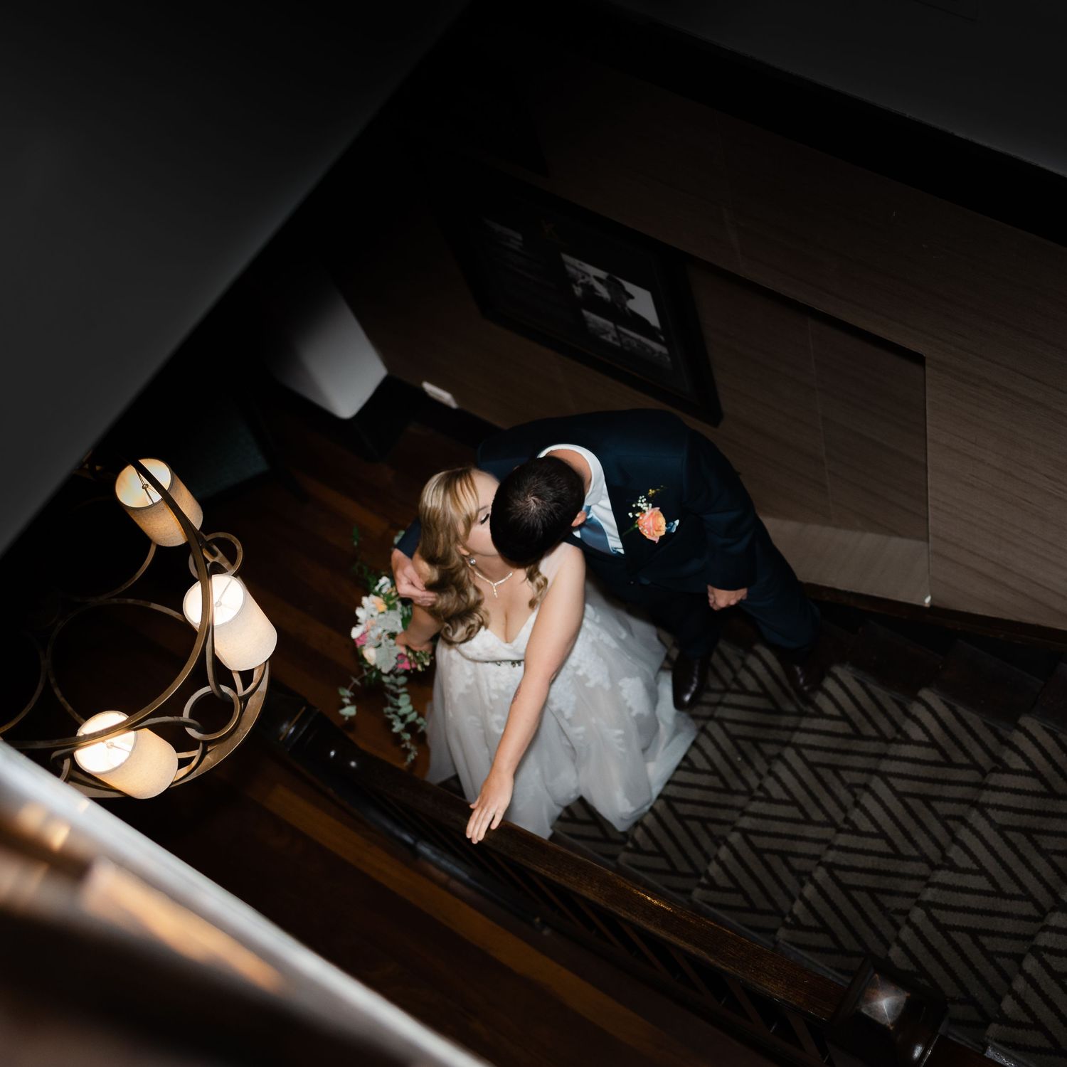 Aerial view of a bride and groom sharing a kiss on a staircase, bride holding a bouquet, warm ambient lighting.