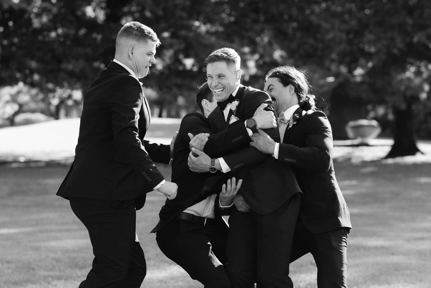 A group of people in formal black attire dancing together outdoors in a playful manner.
