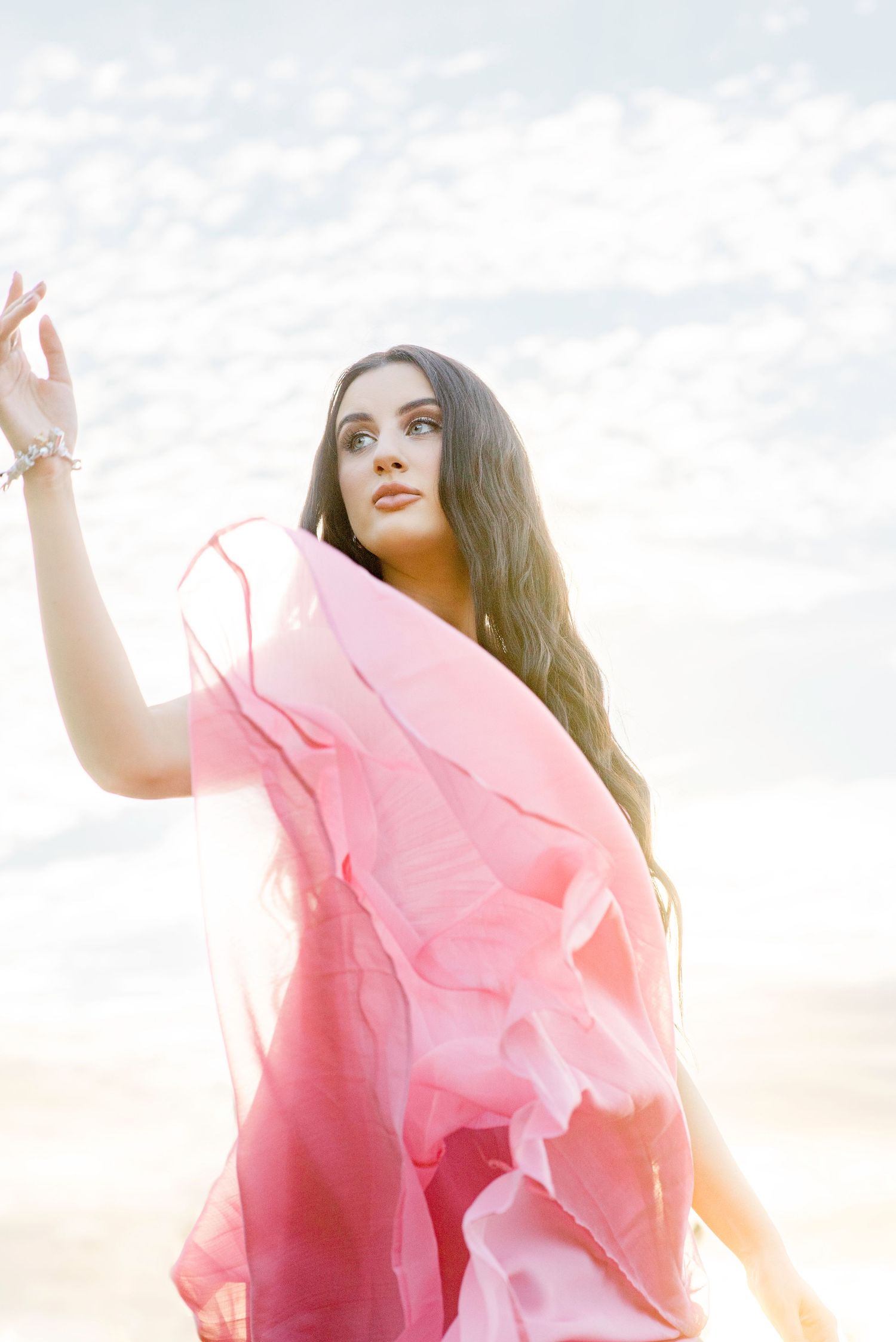 A woman in a flowing pink sheer dress poses gracefully on a beach with her arm raised.