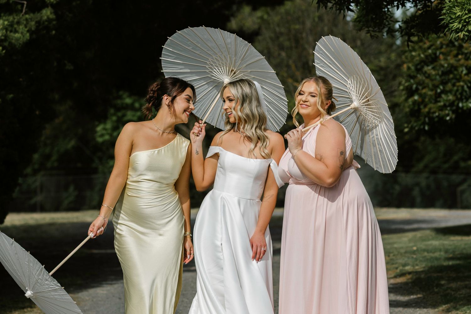 Bride in white gown with two bridesmaids in satin dresses holding white parasols outdoors.