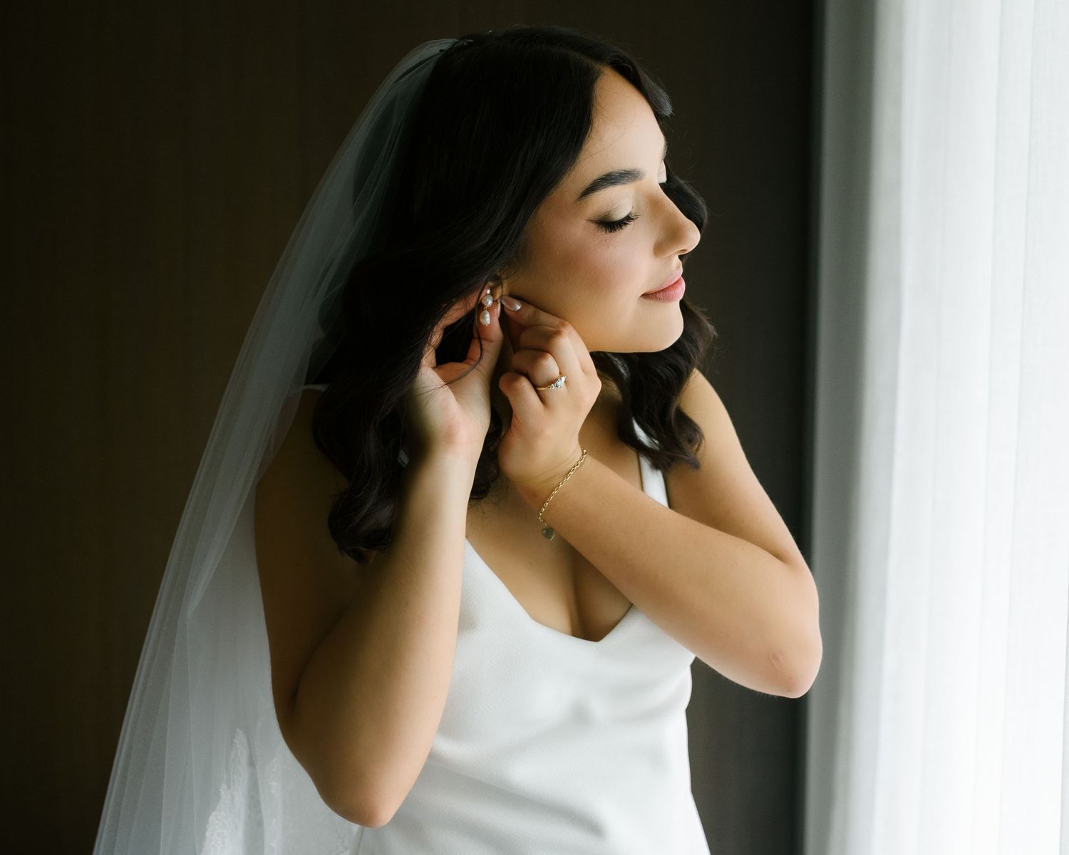 Bride in white gown adjusting earrings with veil, soft natural light, elegant bridal getting ready moment.