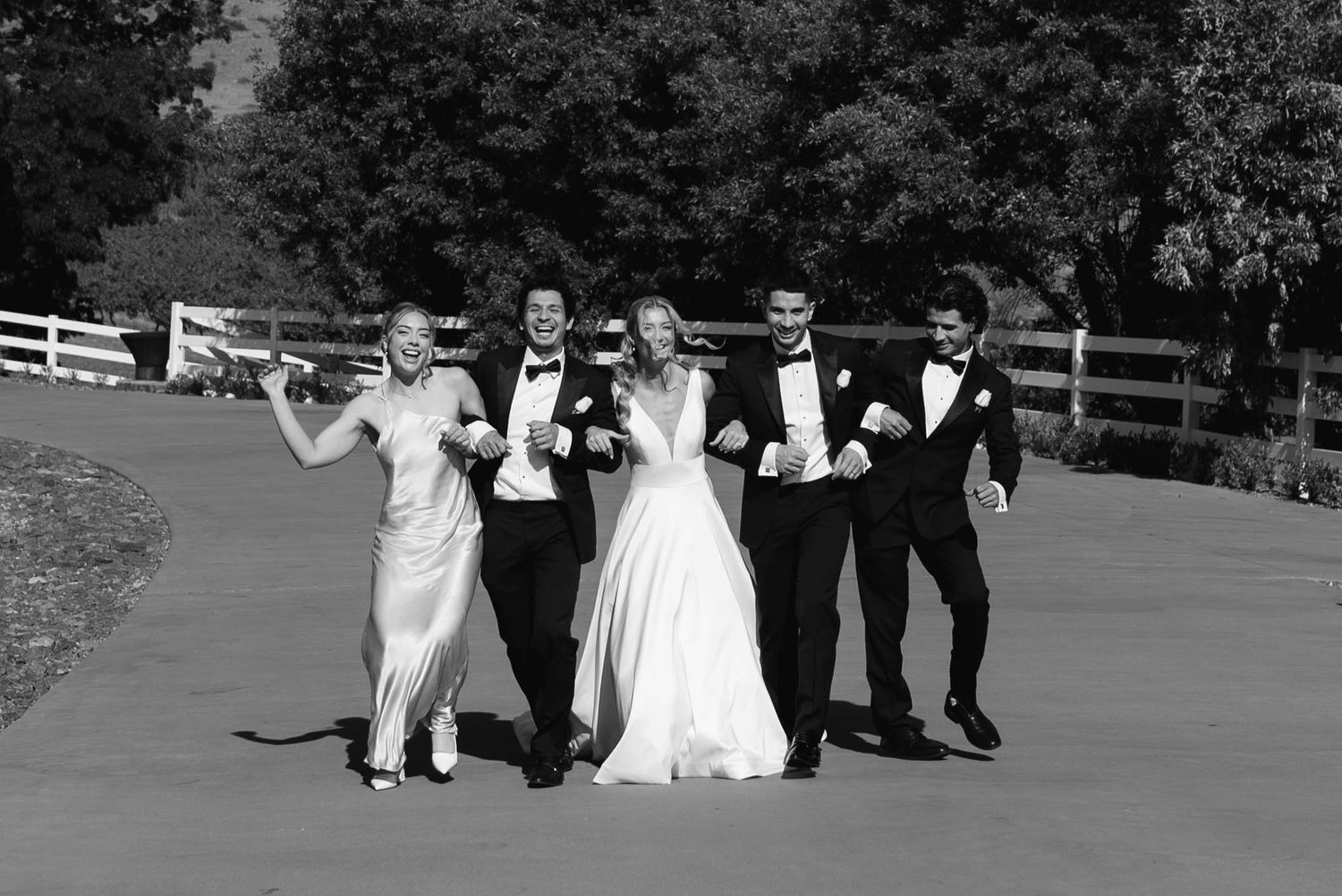 Black and white photo of a bride and groom celebrating with wedding party on a sunny outdoor path.