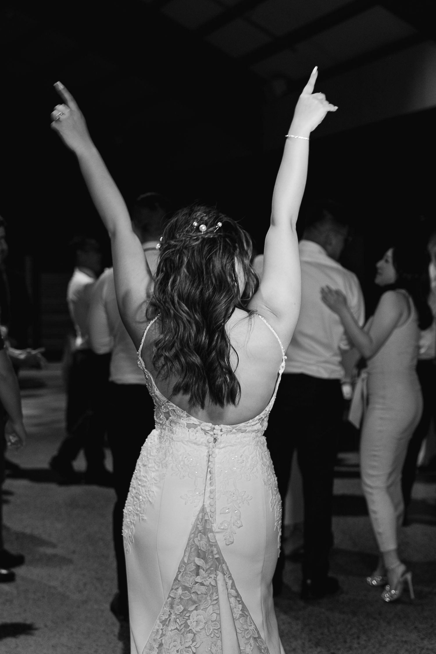 A person in a white lace dress dances with their arms raised on a dark dance floor at a celebration.