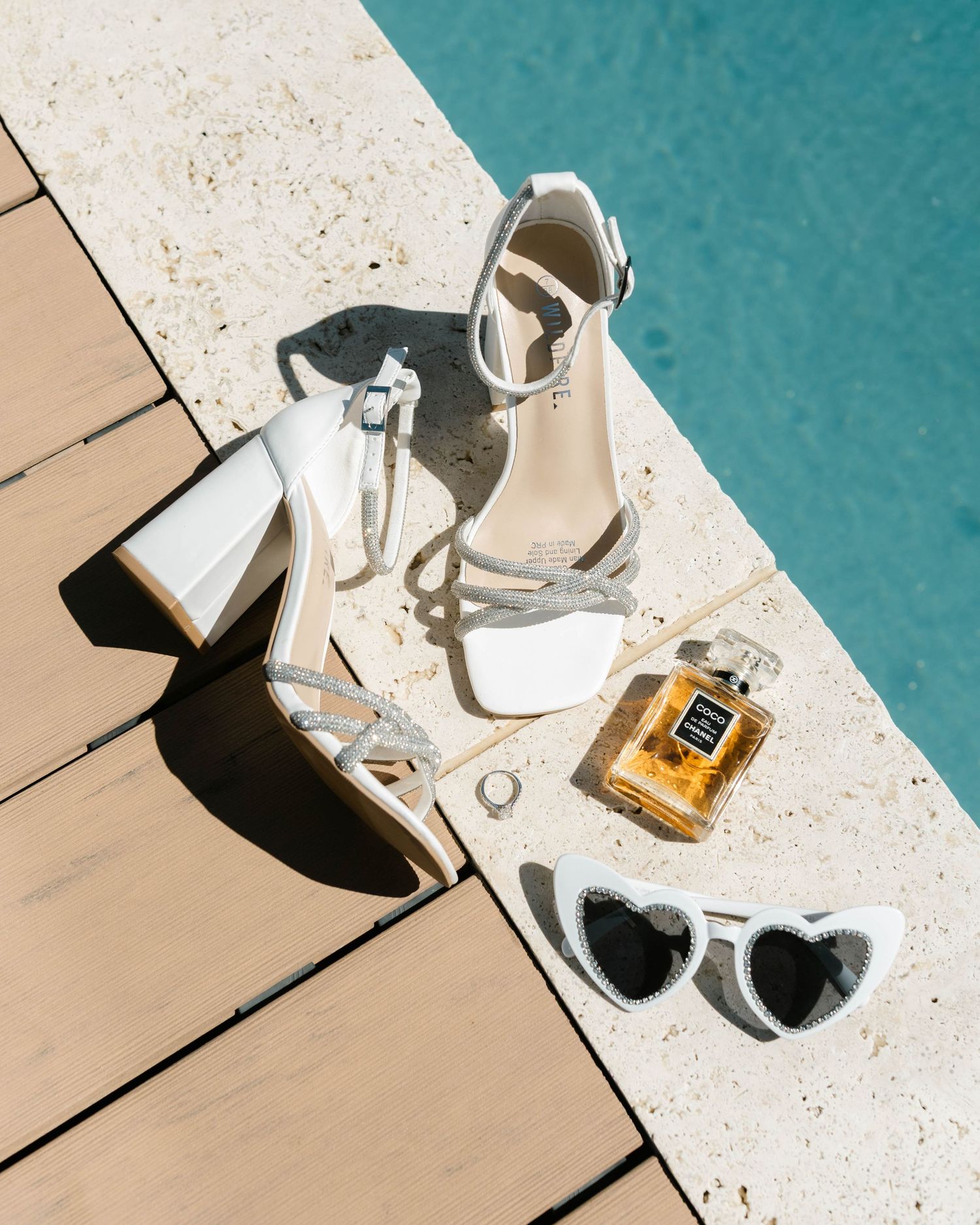 White bridal shoes and perfume bottle arranged on a wooden deck near turquoise water.