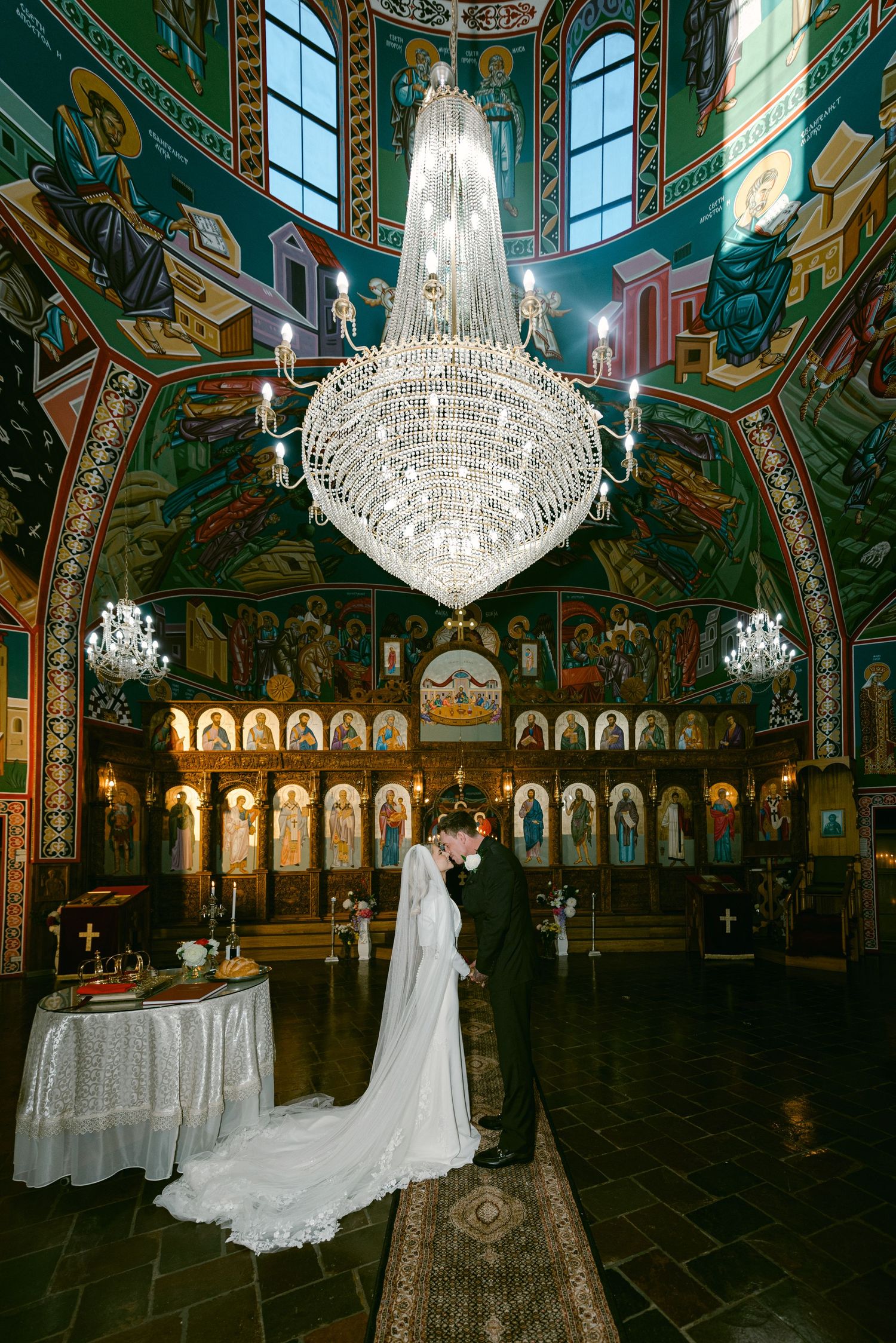 Bride and groom stand beneath crystal chandelier in ornate Orthodox church with colorful Byzantine frescoes and golden iconostasis.