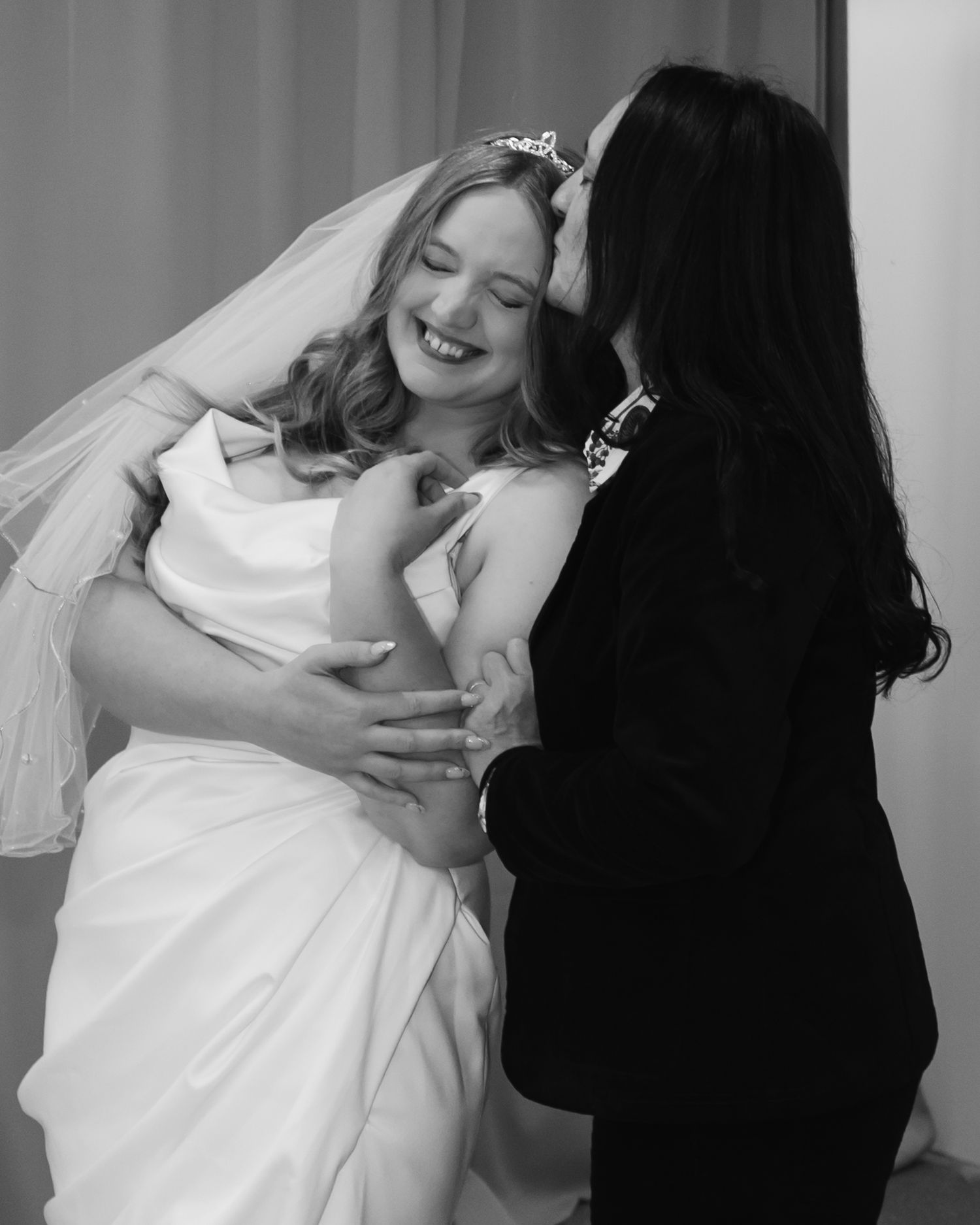 Black and white photo of a smiling bride in veil and tiara being embraced and kissed on the cheek at her wedding.