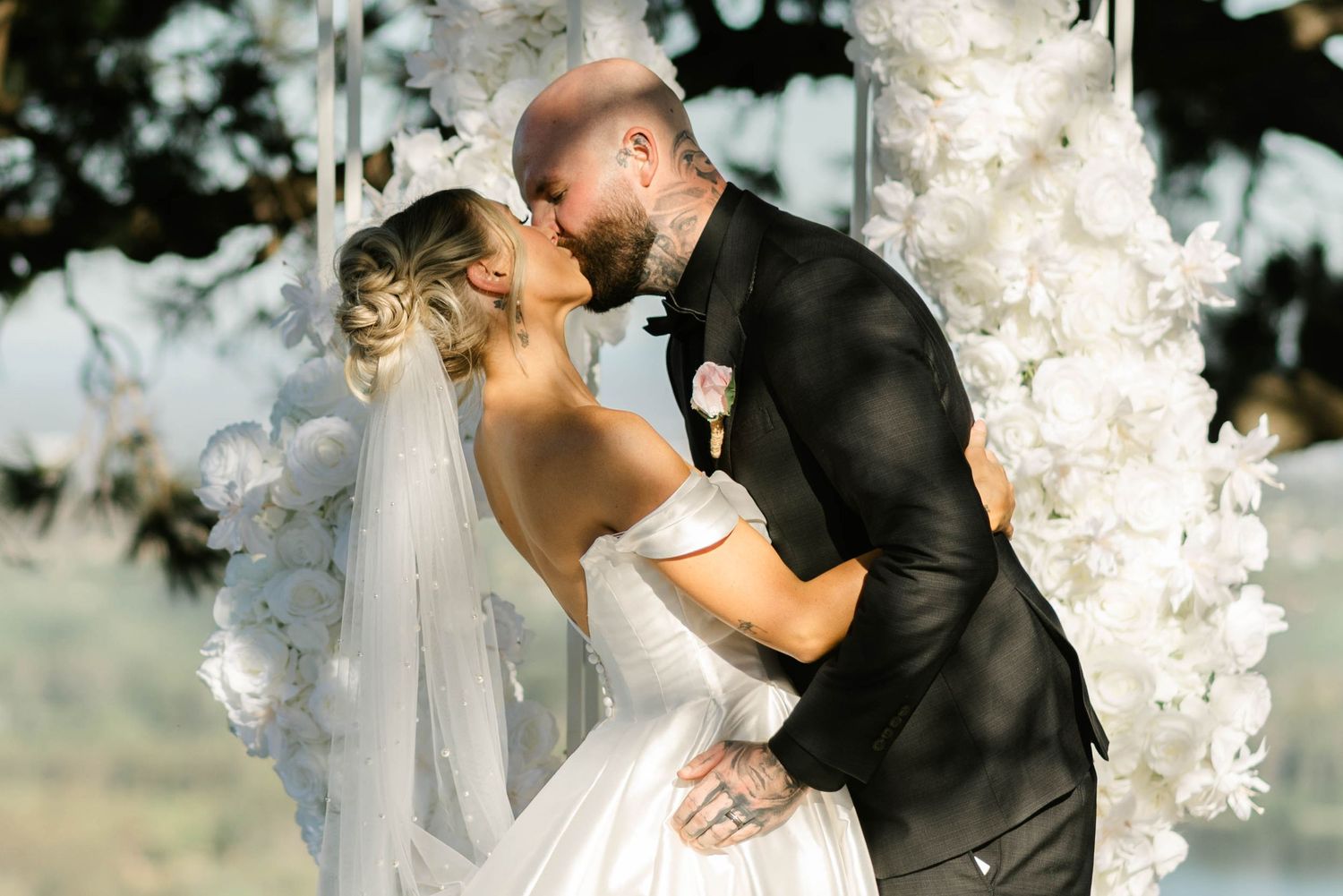 Romantic wedding couple shares a kiss under a white floral arch during an outdoor ceremony.