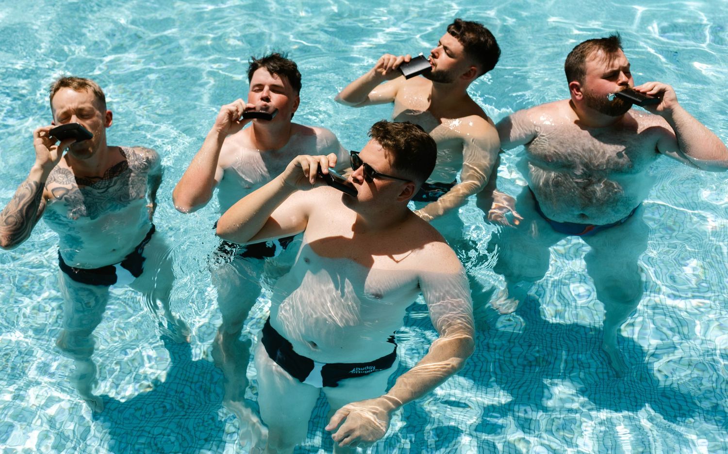 Five shirtless men standing in a swimming pool, drinking beverages together on a sunny day.